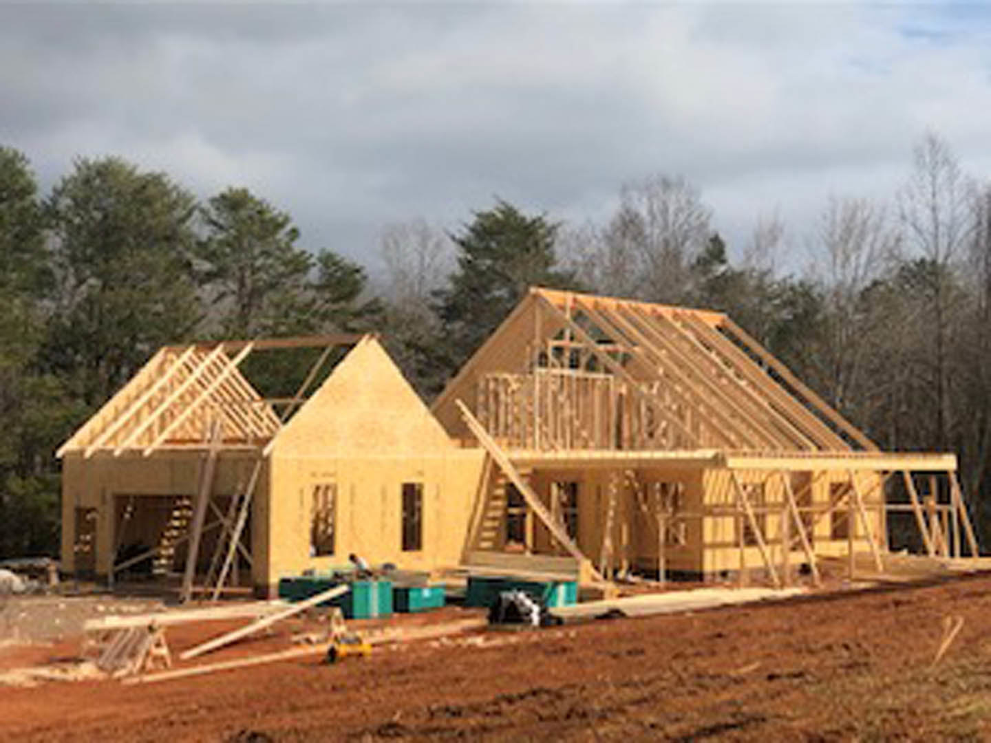 Framed wooden house under construction on dirt lot, surrounded by trees and cloudy sky