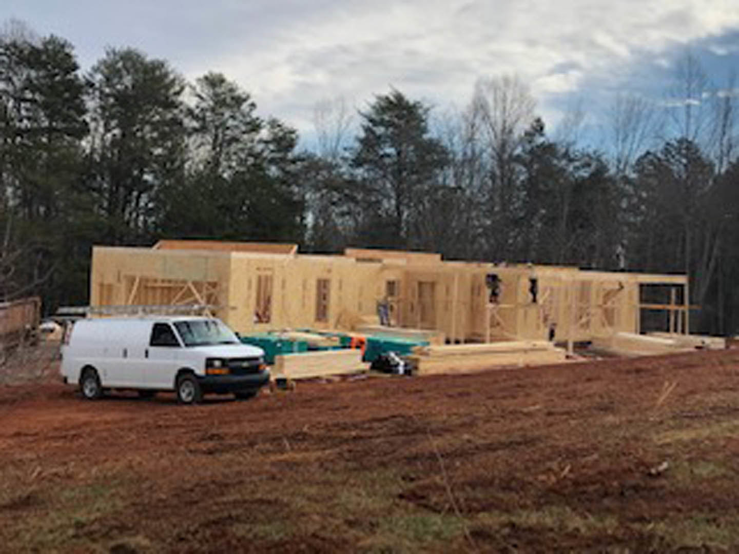 Modern home under construction with exposed framing, surrounded by trees, white van parked on gravel driveway, cloudy sky overhead