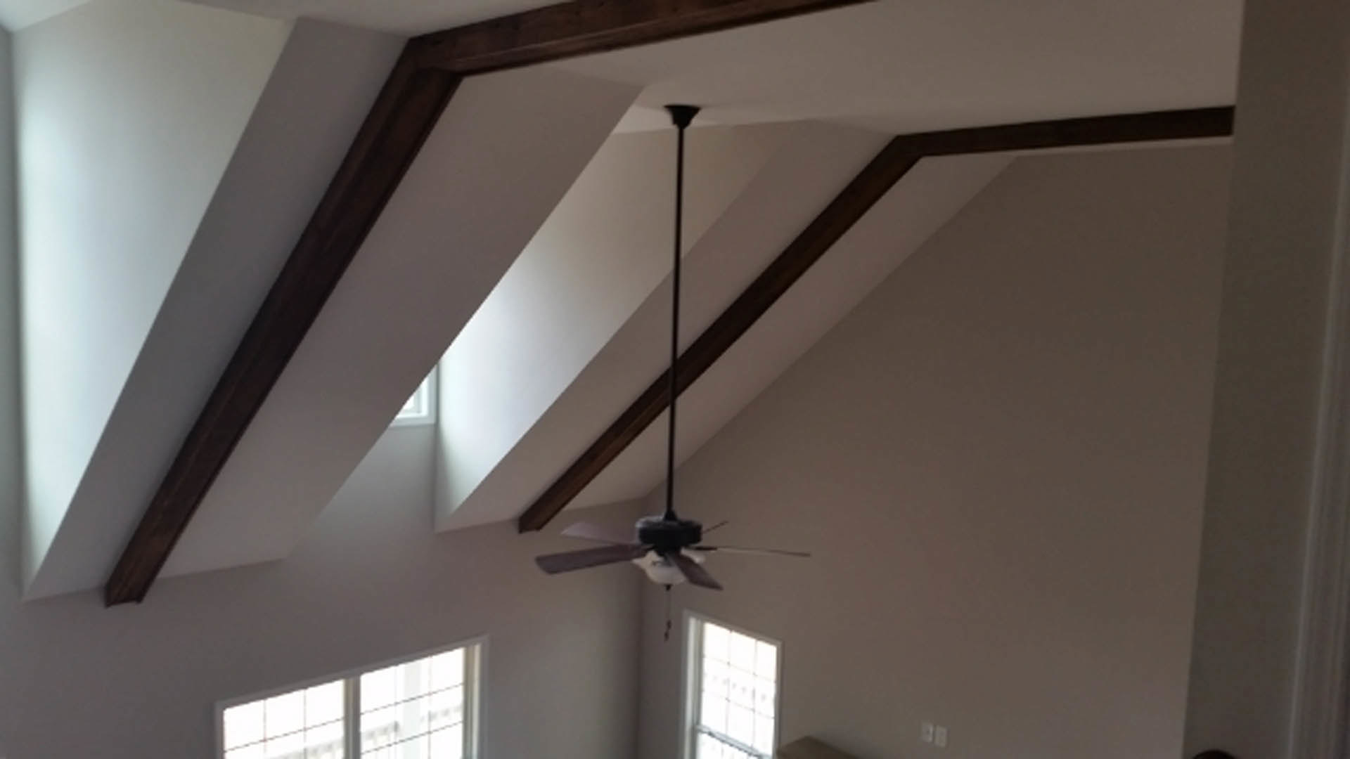Ceiling fan with light fixture mounted on beamed ceiling, white framed window letting daylight into plaster-walled room