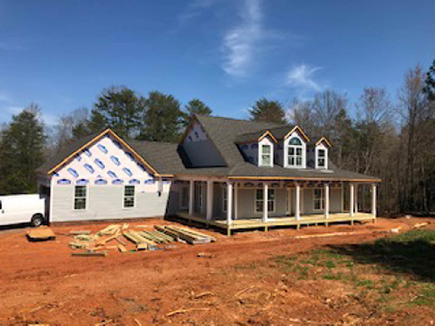 Partially built house with triangular roof, exposed framing, red dirt construction site, scattered building materials, cloudy sky, and surrounding trees