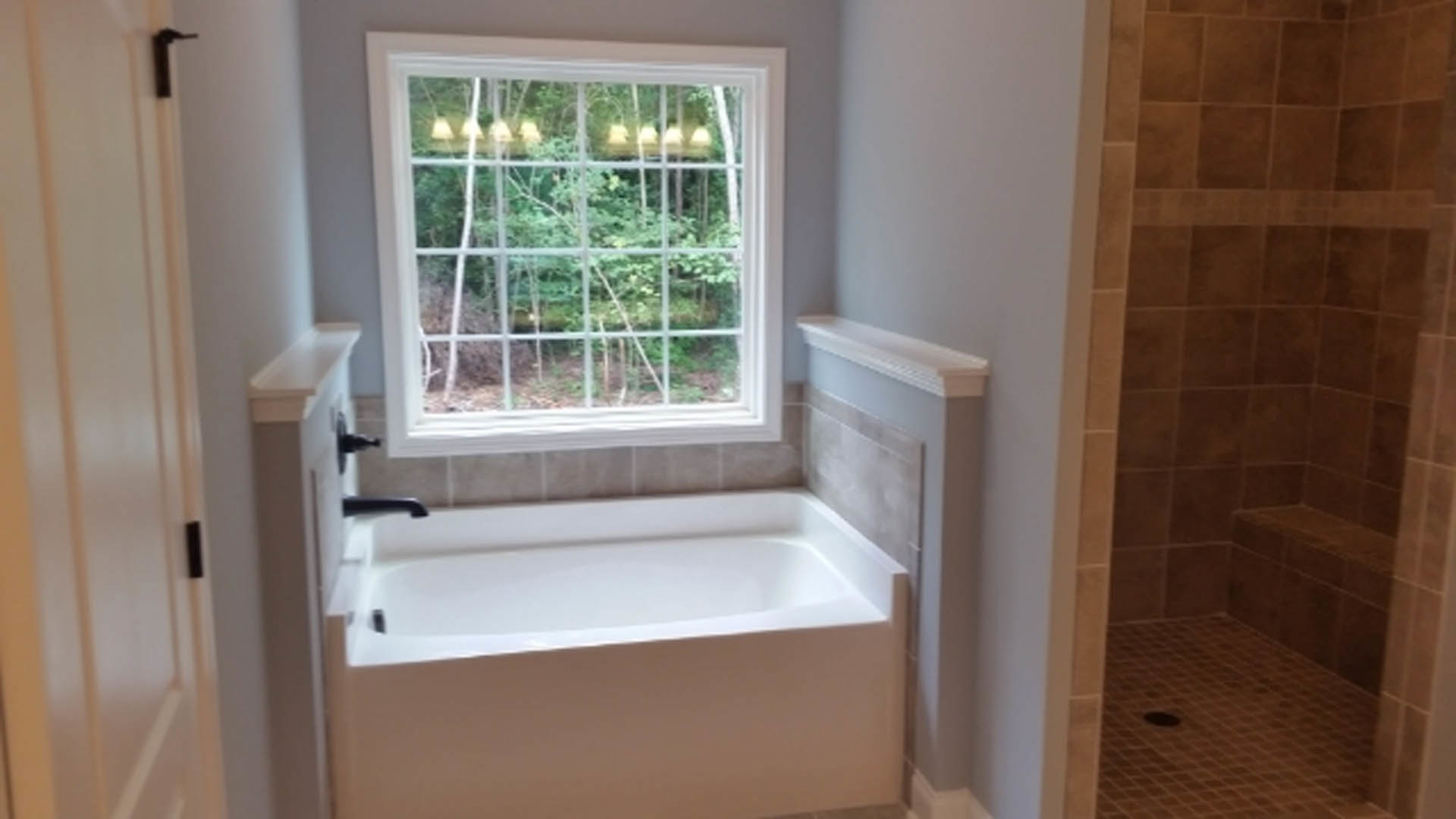 White freestanding bathtub beneath a large window, surrounded by light gray tile walls and chrome fixtures in a modern bathroom.