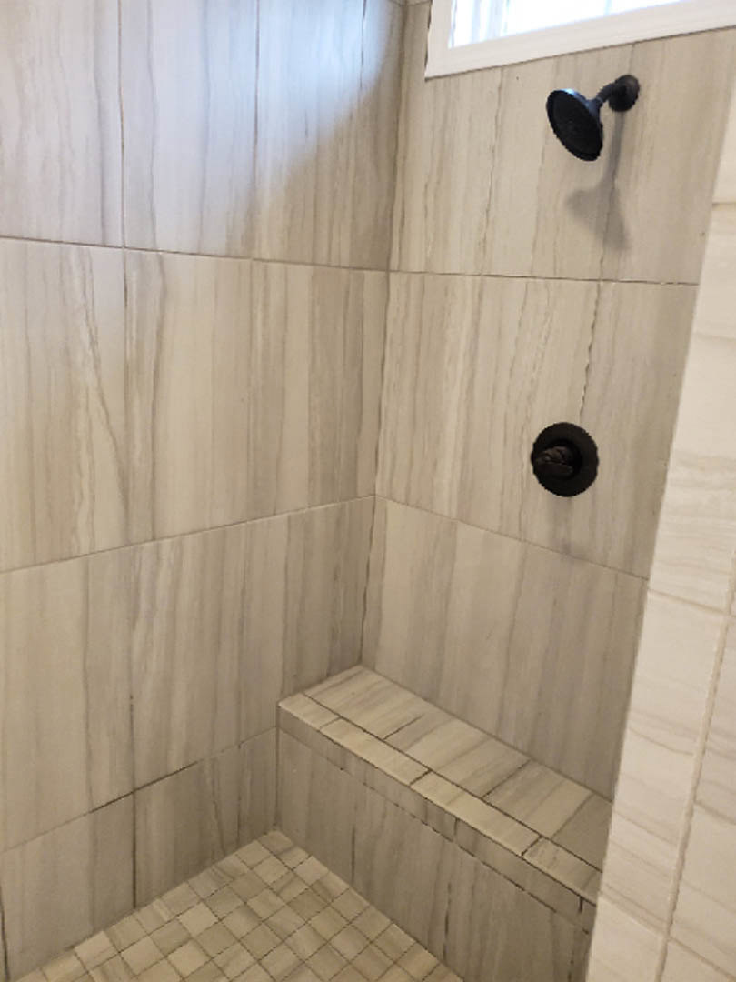 Walk-in shower with gray tile floor, black shower head mounted on tiled wall, partial view of blurry face through opening in wall.