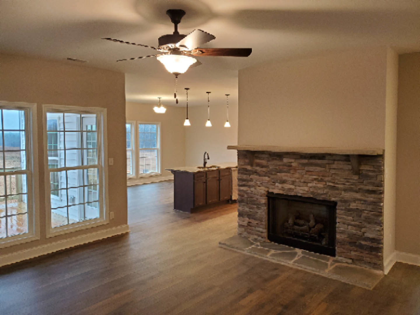 Living room with stone fireplace, lit fire, wood flooring, ceiling fan with light, large window showing exterior white house and greenery