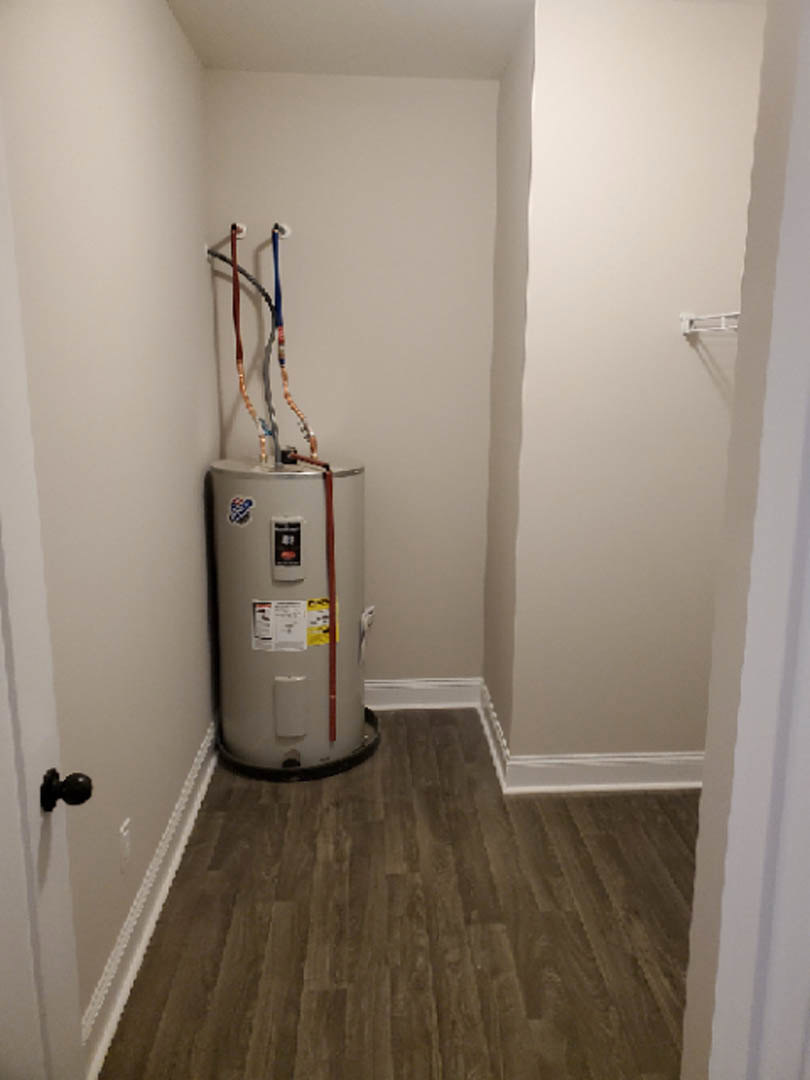 White water heater with visible pipes against a plaster wall in a utility room, wood flooring, fire extinguisher, and refrigerator nearby