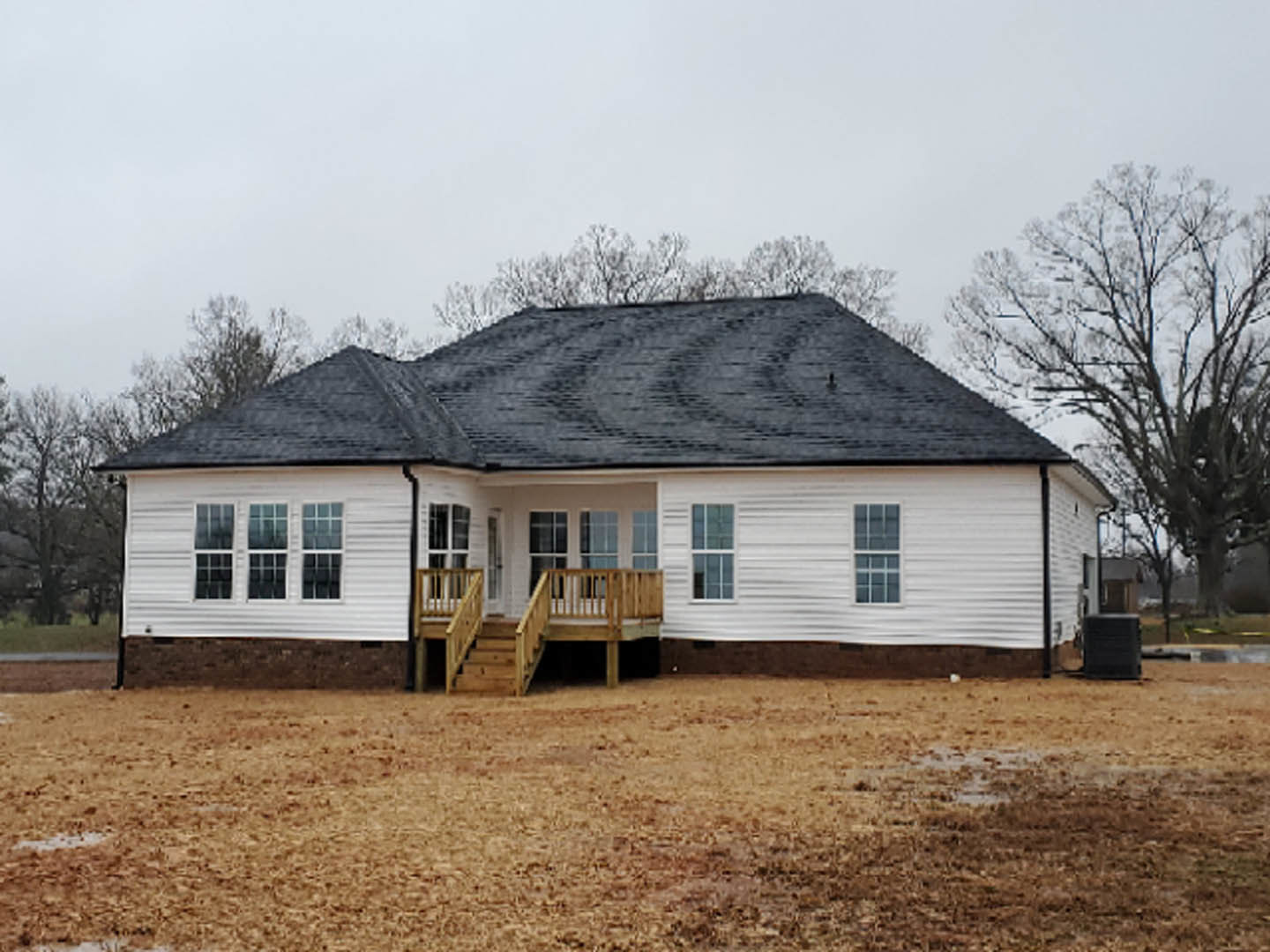 Two-story home with light siding, wooden deck and railing, staircase leading to front door, grassy yard, mature trees in background, large window on main level.