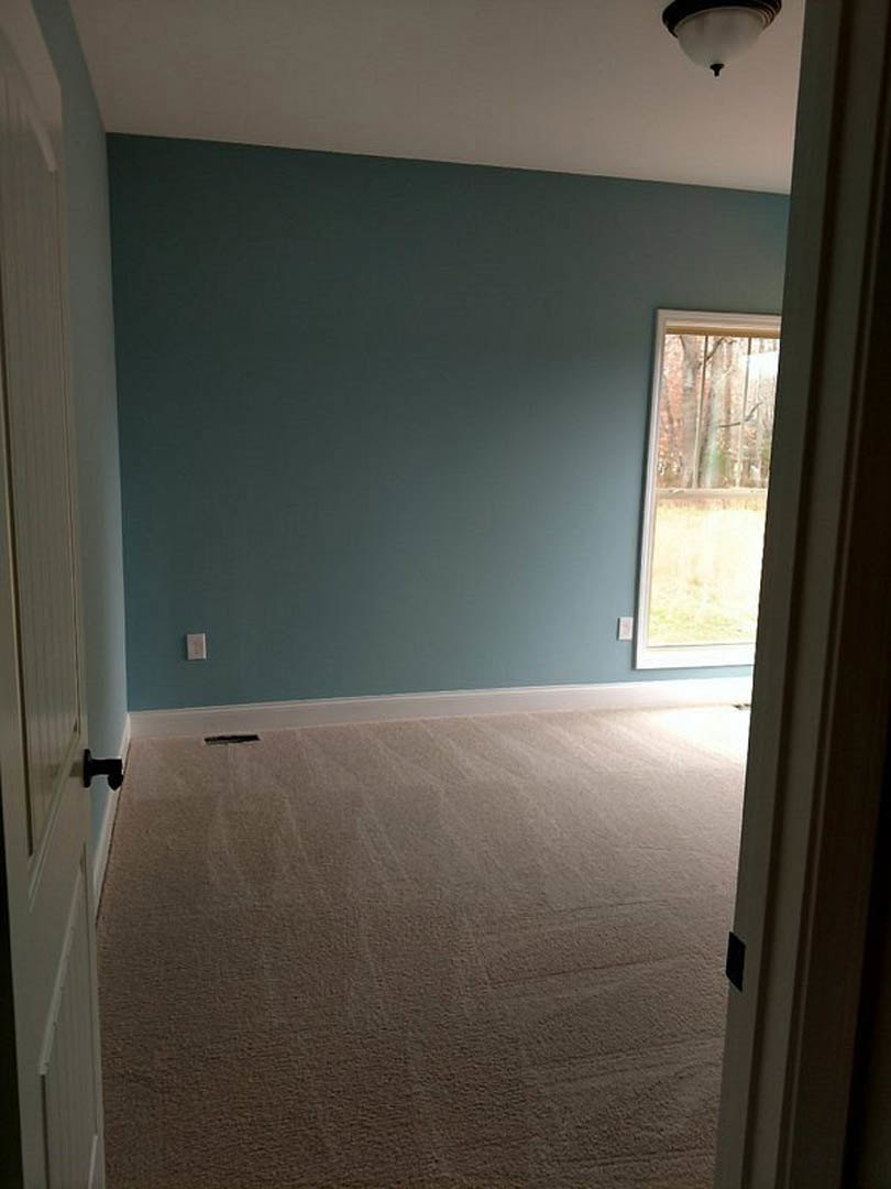 Blue accent wall, carpeted floor with floor vent, large window showing leafy trees, ceiling light fixture, white trim and baseboards