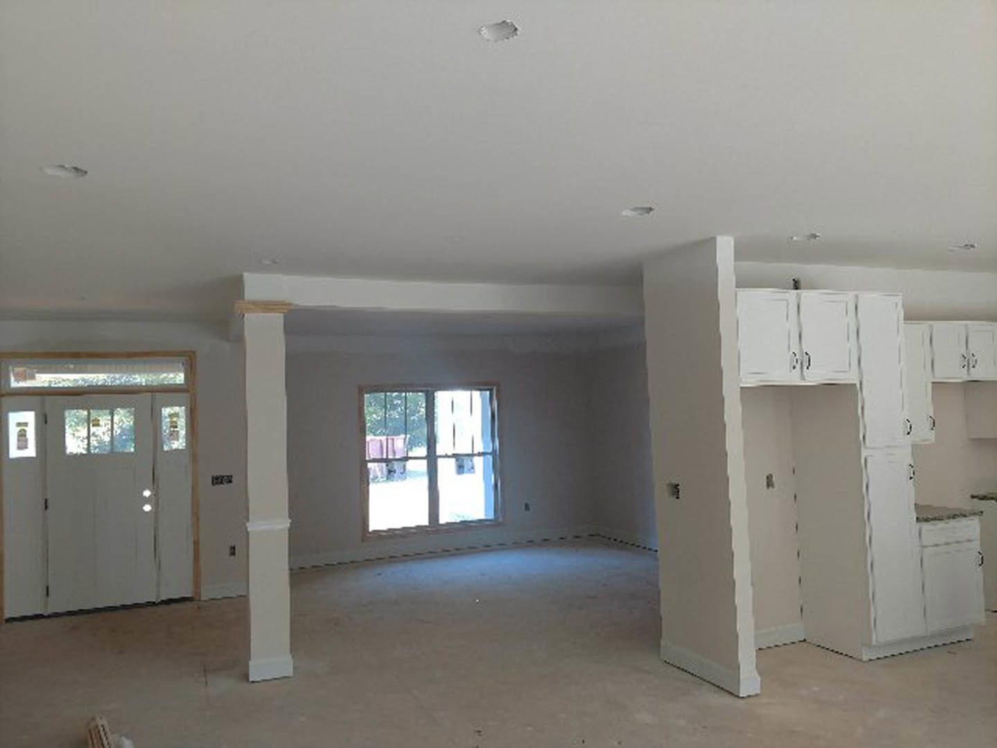 Open room featuring white walls, white columns with black and white bases, white cabinetry in the kitchen area, white door with glass panes, window overlooking fence and trees, and