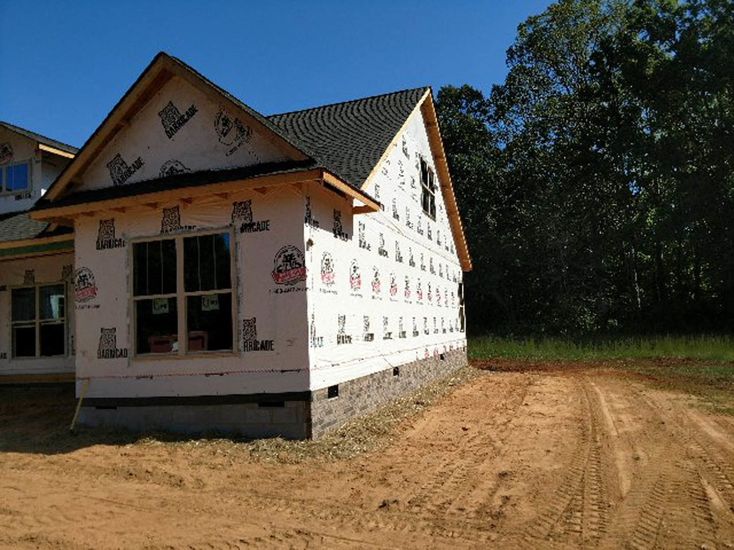 Wood-framed house under construction with exposed sheathing, dirt driveway showing tire tracks, tall trees in background, blue sky overhead, window displaying construction signs