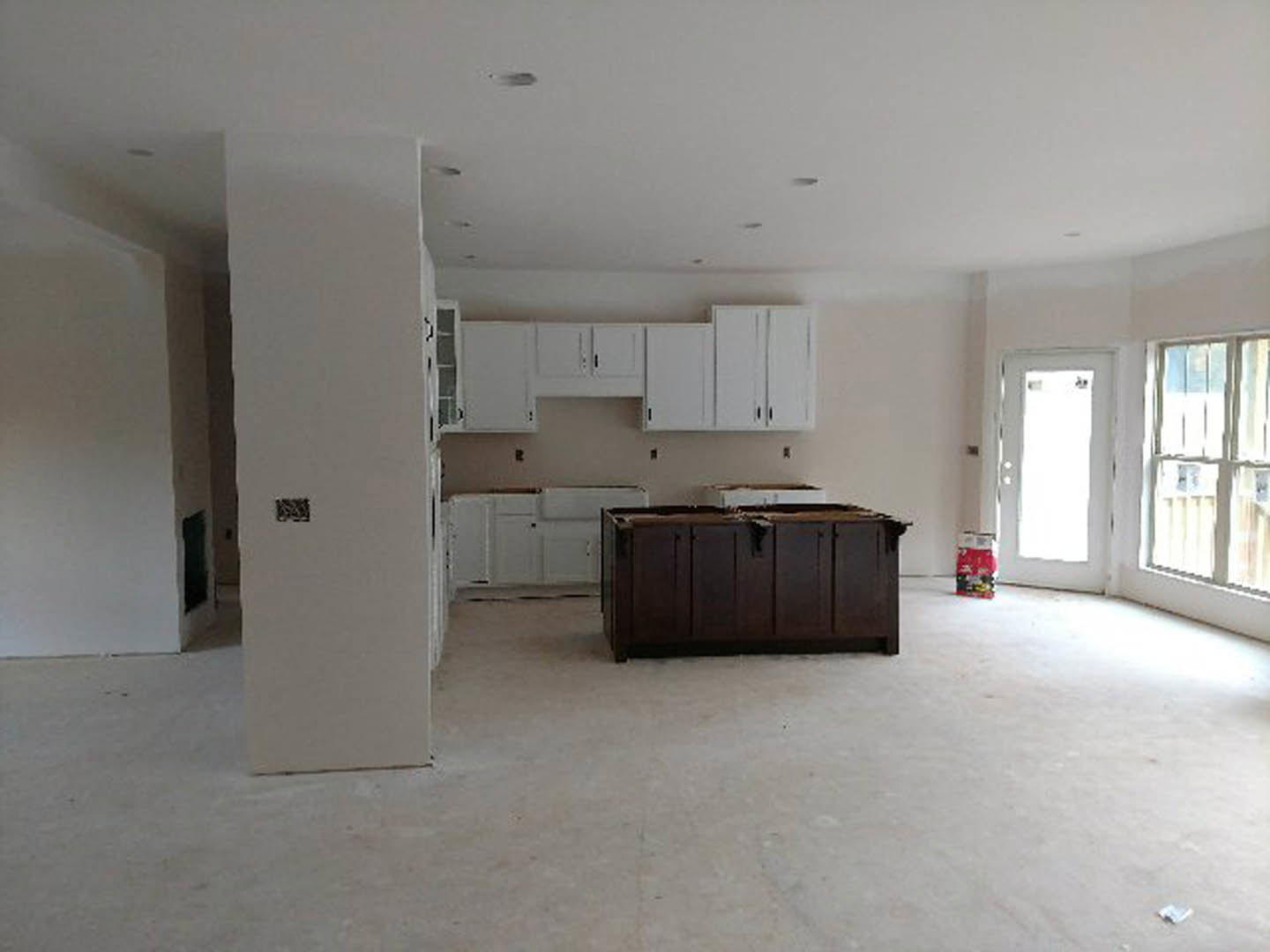 Dark wood lower cabinets with a damaged countertop, white upper cabinets, white-framed window, white door illuminated by bright light, red bag with white text on the floor, plaster