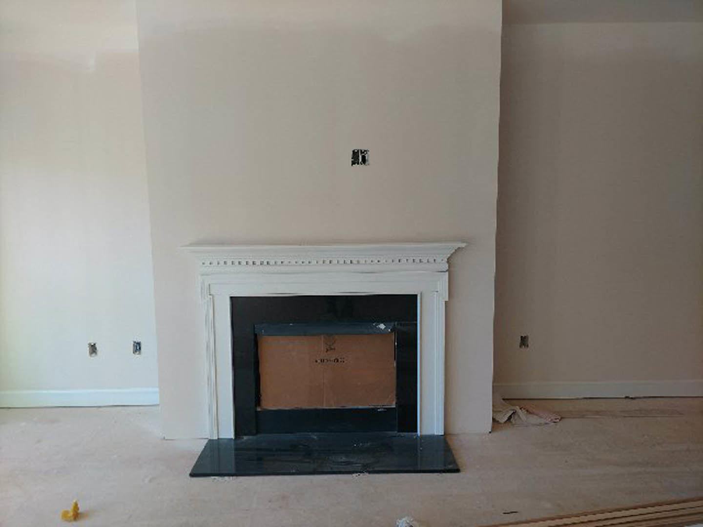 Modern living room with white fireplace surround, wood-burning stove, cardboard box on hearth, yellow object on white mantel shelf, light-colored flooring, and neutral walls