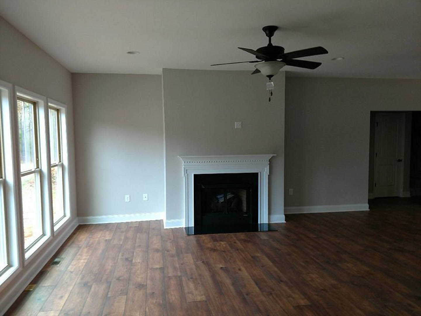 Living room with wood flooring, white-framed fireplace, ceiling fan with light, white door with black handle, and window.