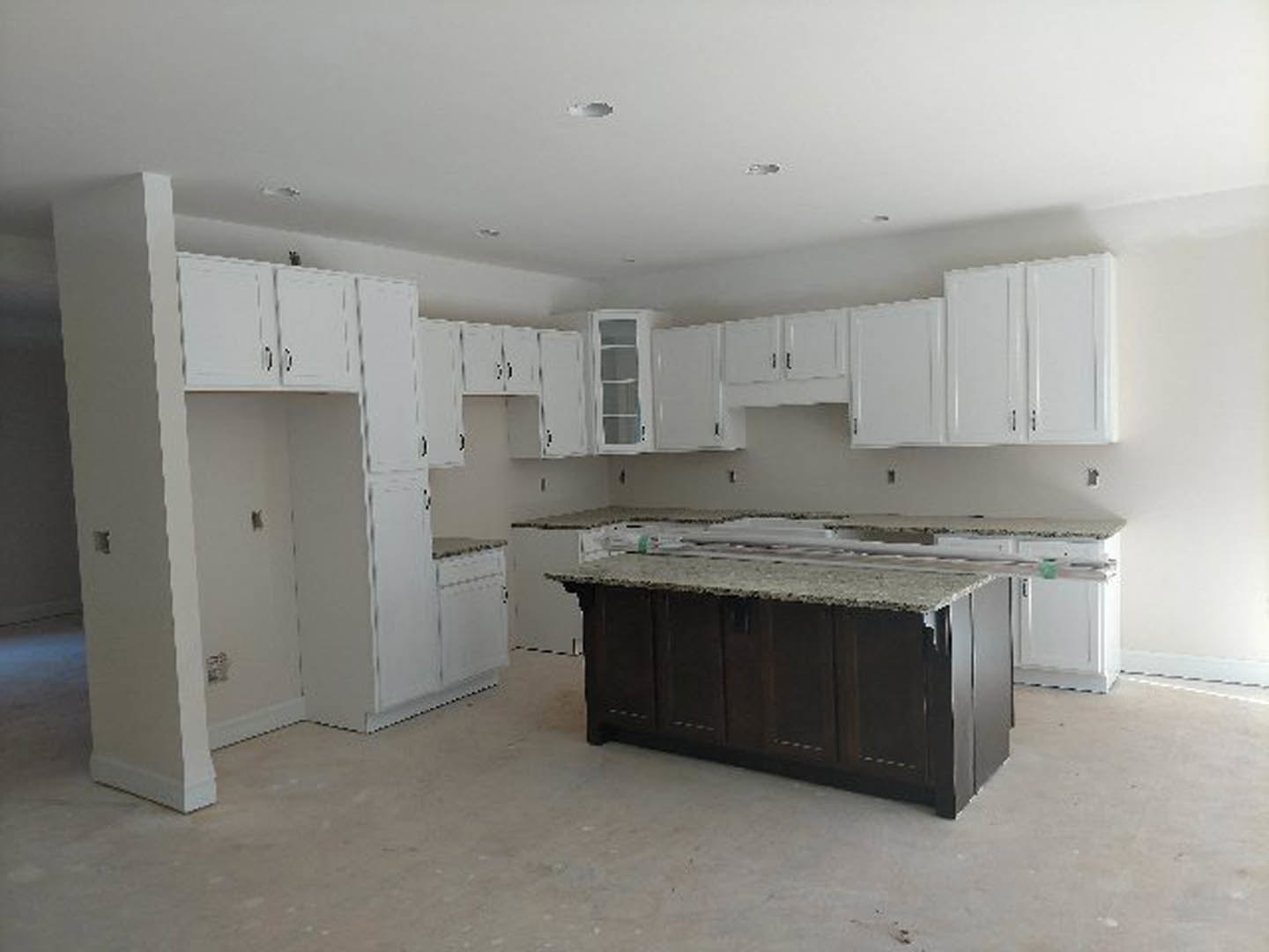 White kitchen cabinets with brushed metal hardware, dark wood island base, light stone countertop, stainless steel sink, white-framed window, concrete slab flooring, and recessed