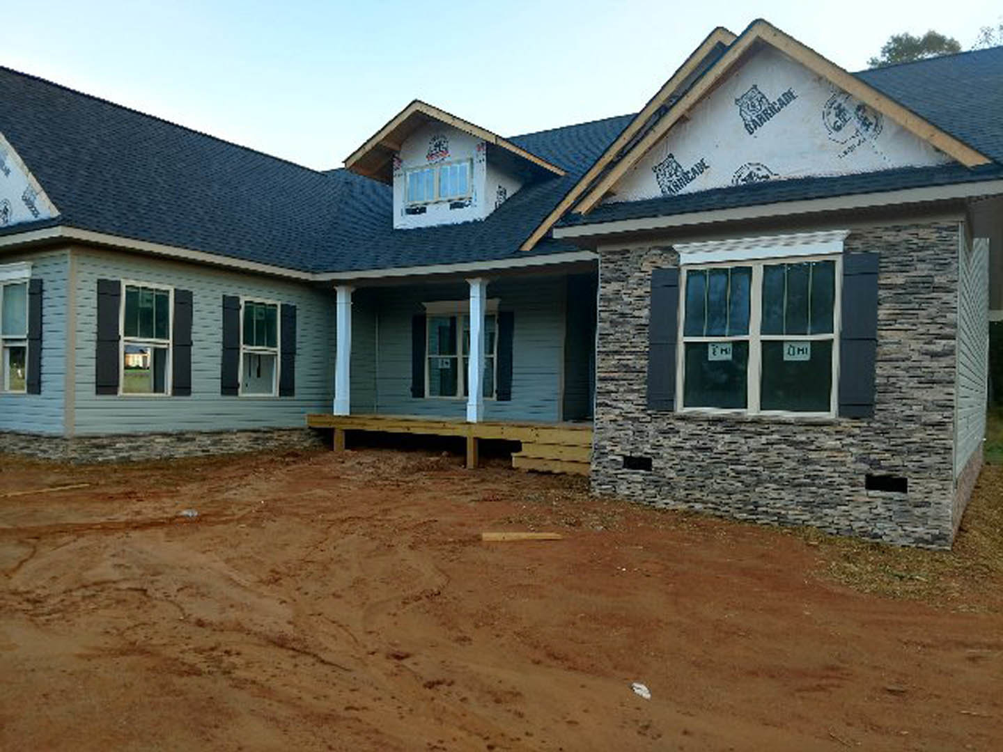 Two-story house with white siding, black roof underlayment, dirt yard, stone retaining wall, wooden platform, and windows with white frames and posted signs