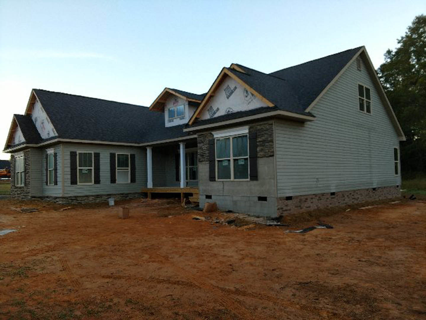 Two-story house under construction with exposed framing, white window frames, concrete foundation, and dirt yard under clear blue sky
