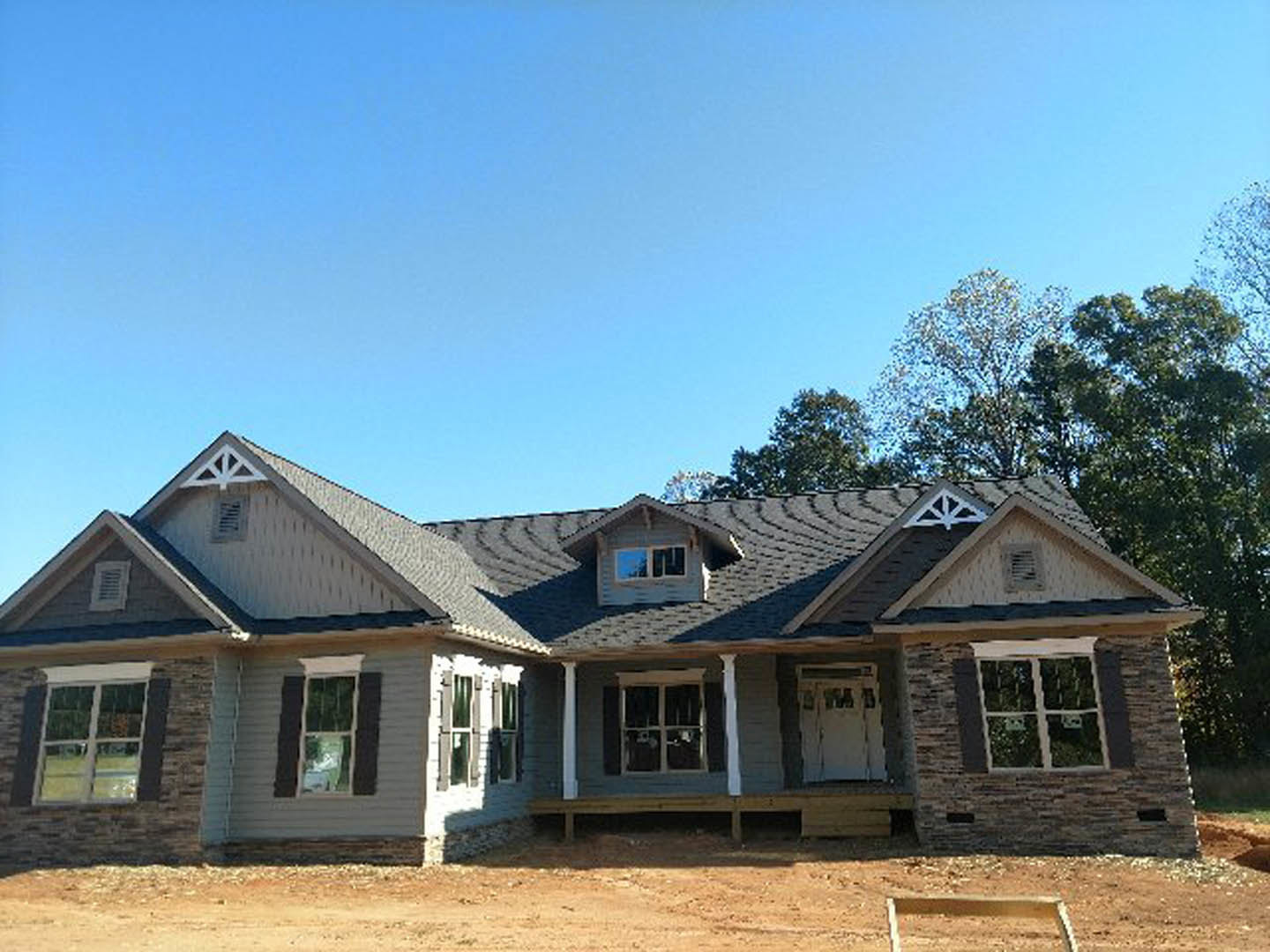 Framed house under construction with exposed wood, white window frames, dirt foreground, and leafy trees in background