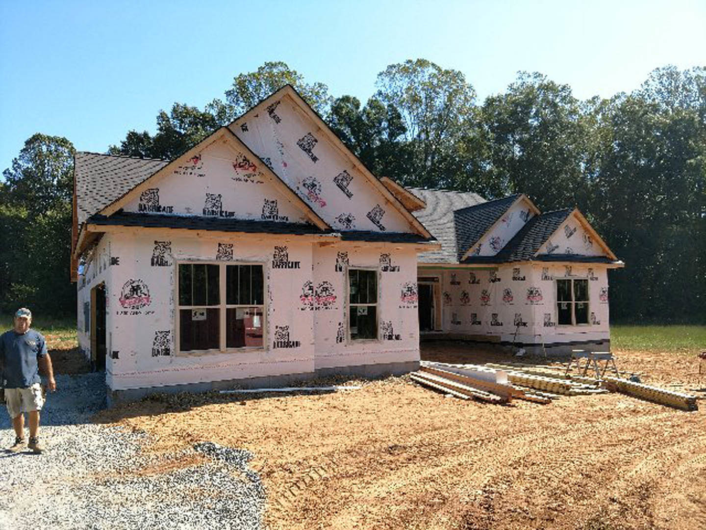 Framed custom home under construction with exposed wood beams, unfinished roof, and surrounding mature trees in the background