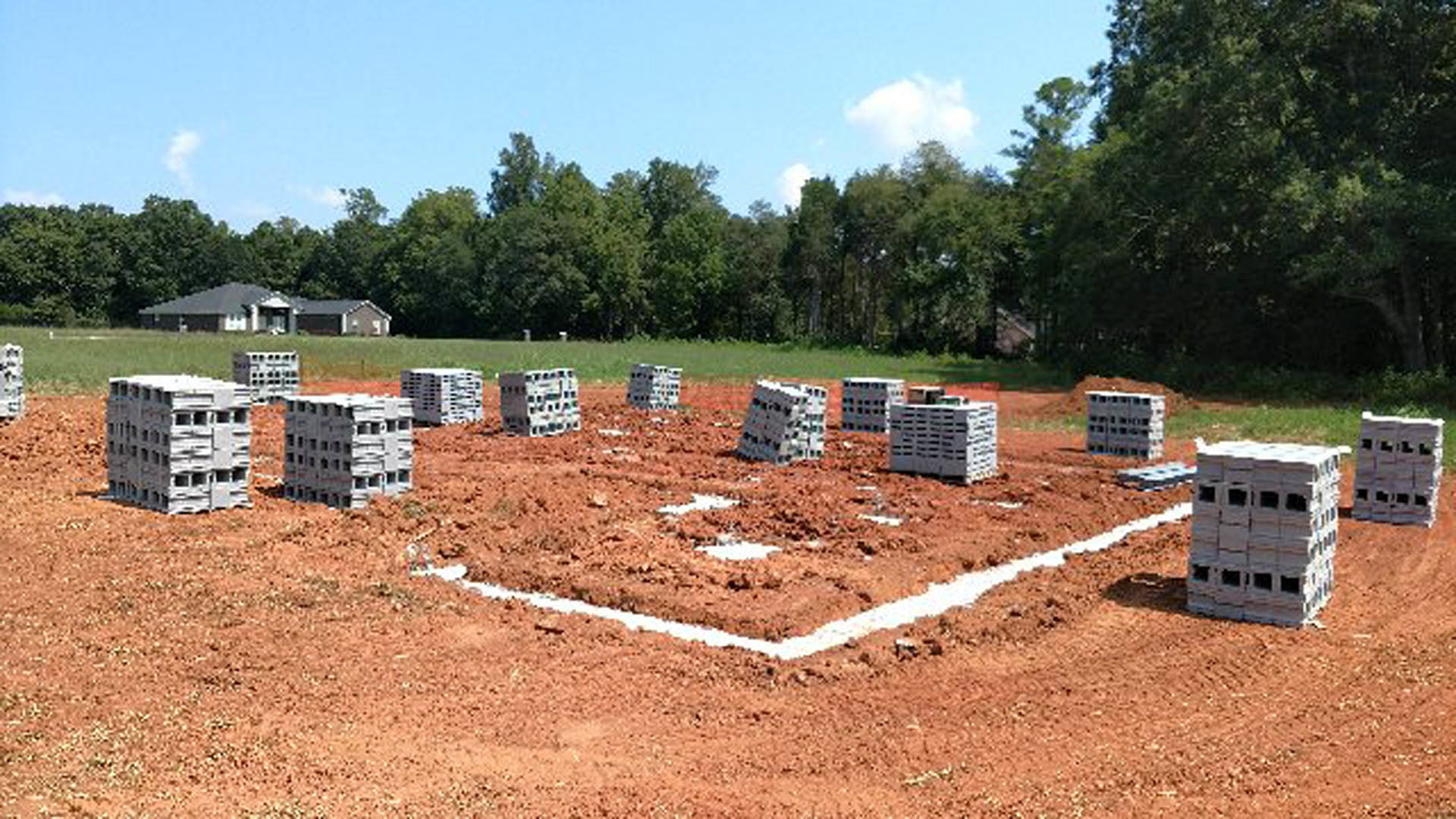 Construction site with stacks of grey crates on bare soil, surrounded by trees and plants under a cloudy sky