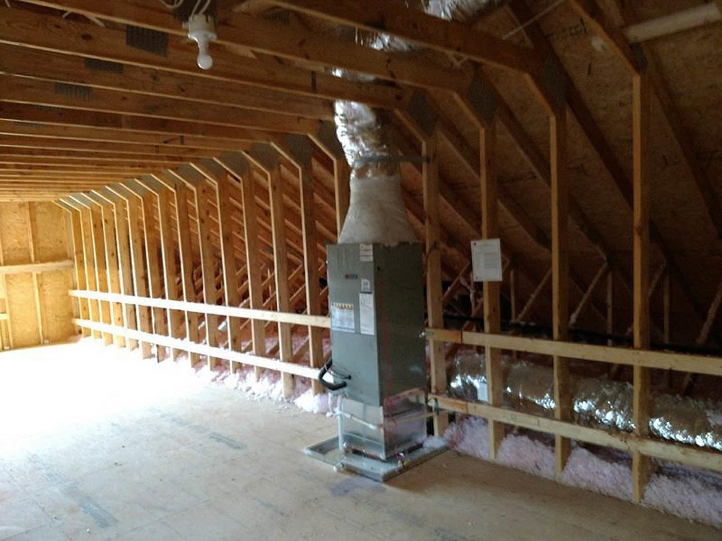 Unfinished attic with exposed wooden beams, metal HVAC box with vent and black handle, white insulation piled against wall, and paper label attached to unit