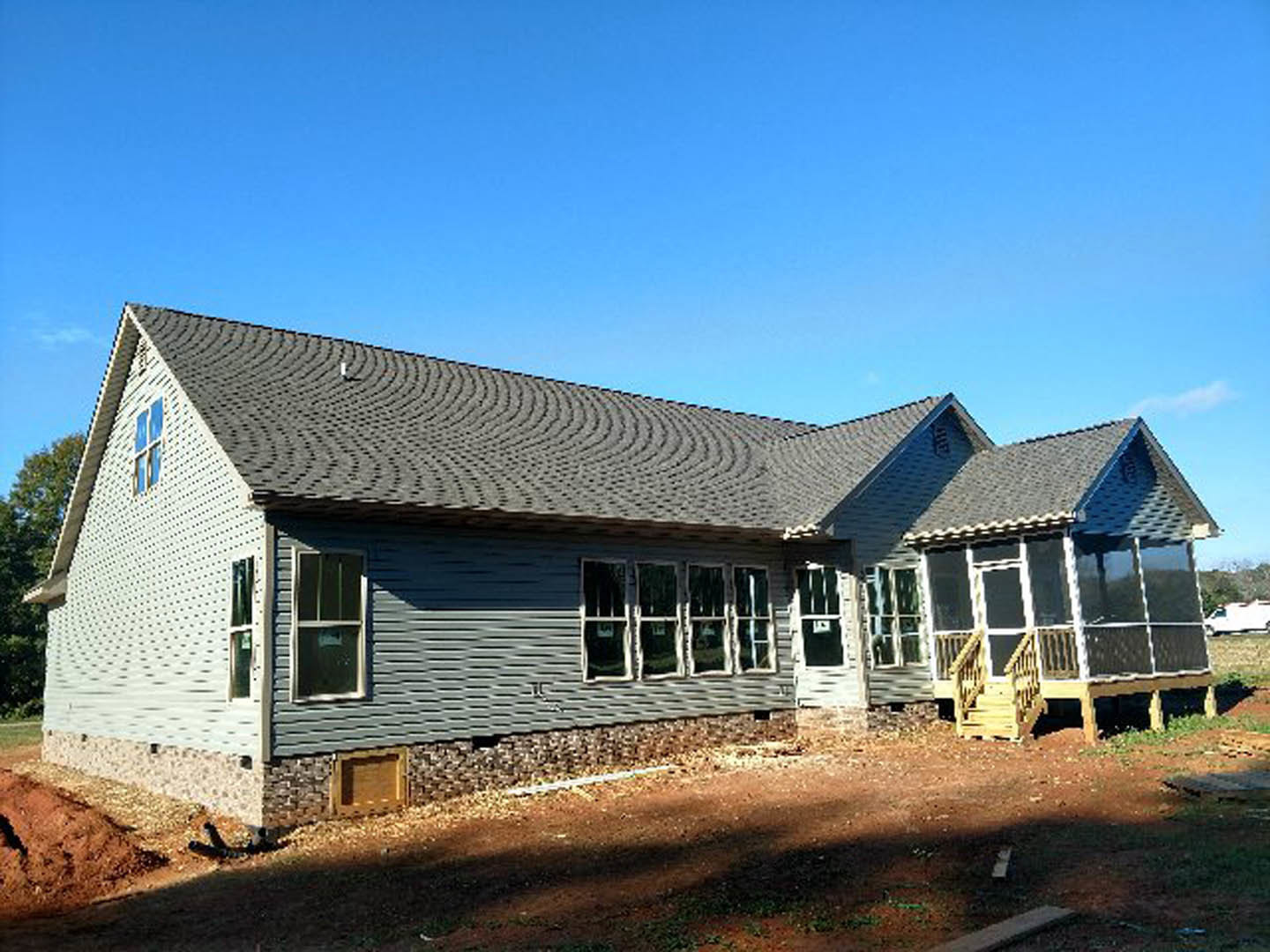 Wood-framed house under construction with covered porch, white and black window frames, exposed sheathing, and blue sky overhead