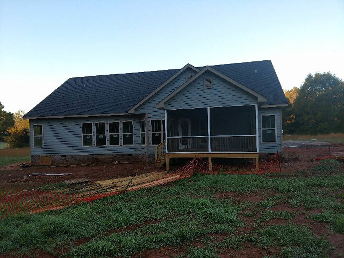 Two-story house under construction with exposed framing, white window frames, covered porch, wooden fence, leafy tree in front yard, grassy lawn, and blue sky overhead