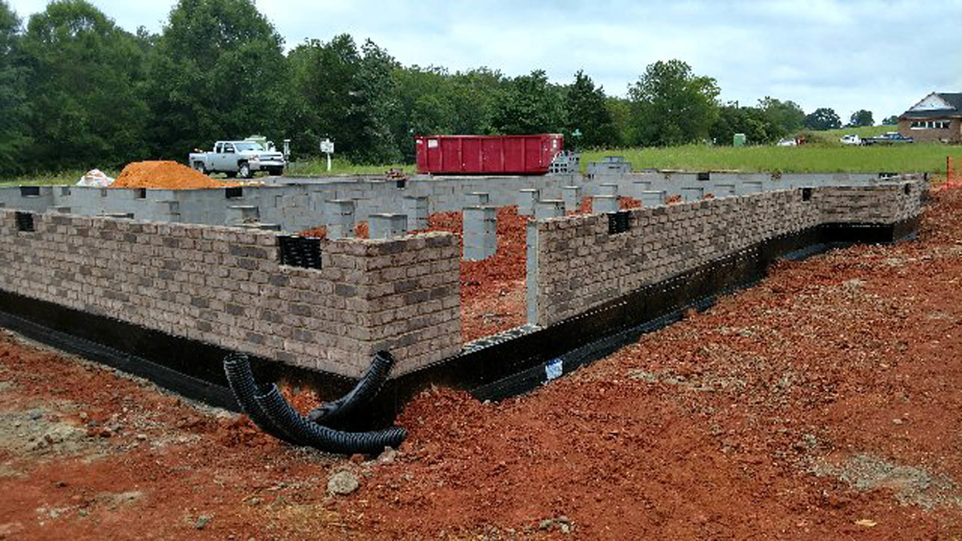 Brick-walled building site with red dirt field, white truck parked nearby, red metal fence partially covered by green leaves, trees and sky in background