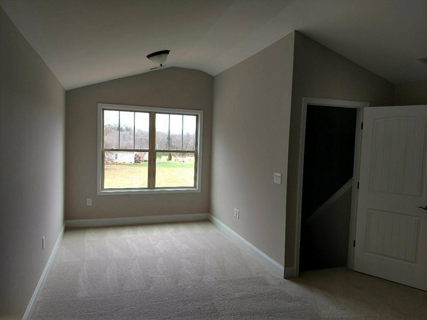 Bedroom with white carpet, large window overlooking grassy field, white walls, white door with black handle, ceiling lamp fixture