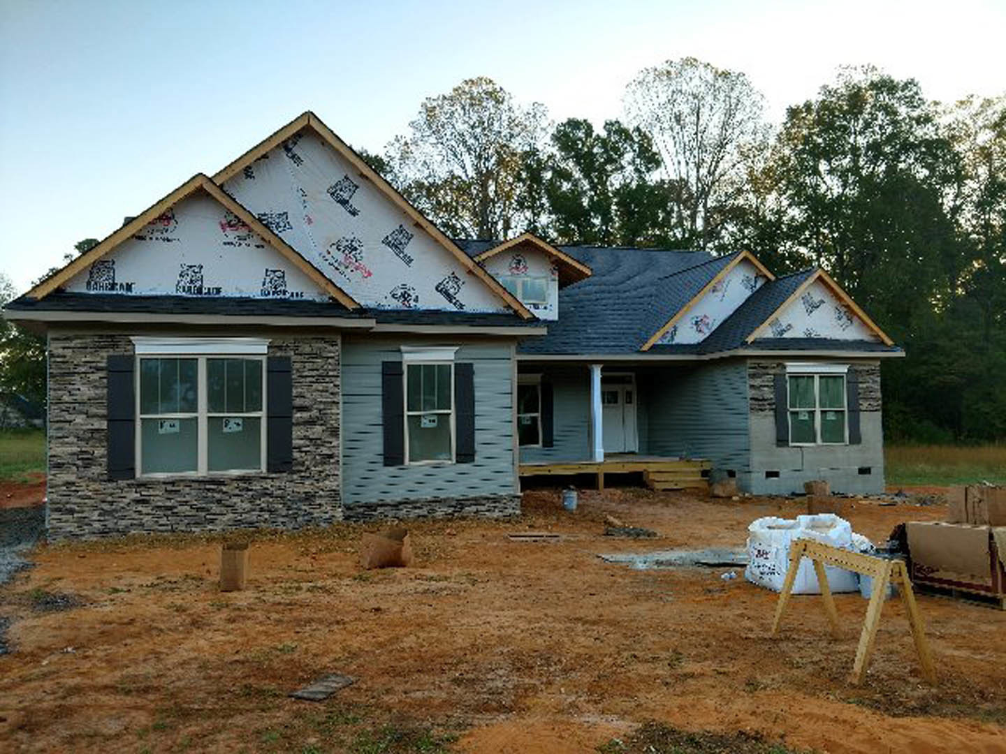 Partially built house framed in wood, surrounded by a temporary fence, with white-framed windows and construction materials scattered on the ground