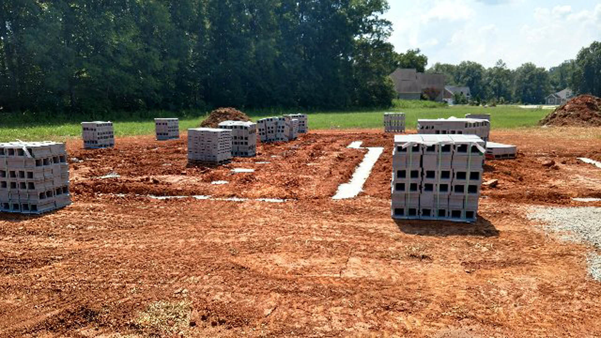 Cement blocks stacked on a dirt lot surrounded by grass and trees, with construction materials scattered across the site.