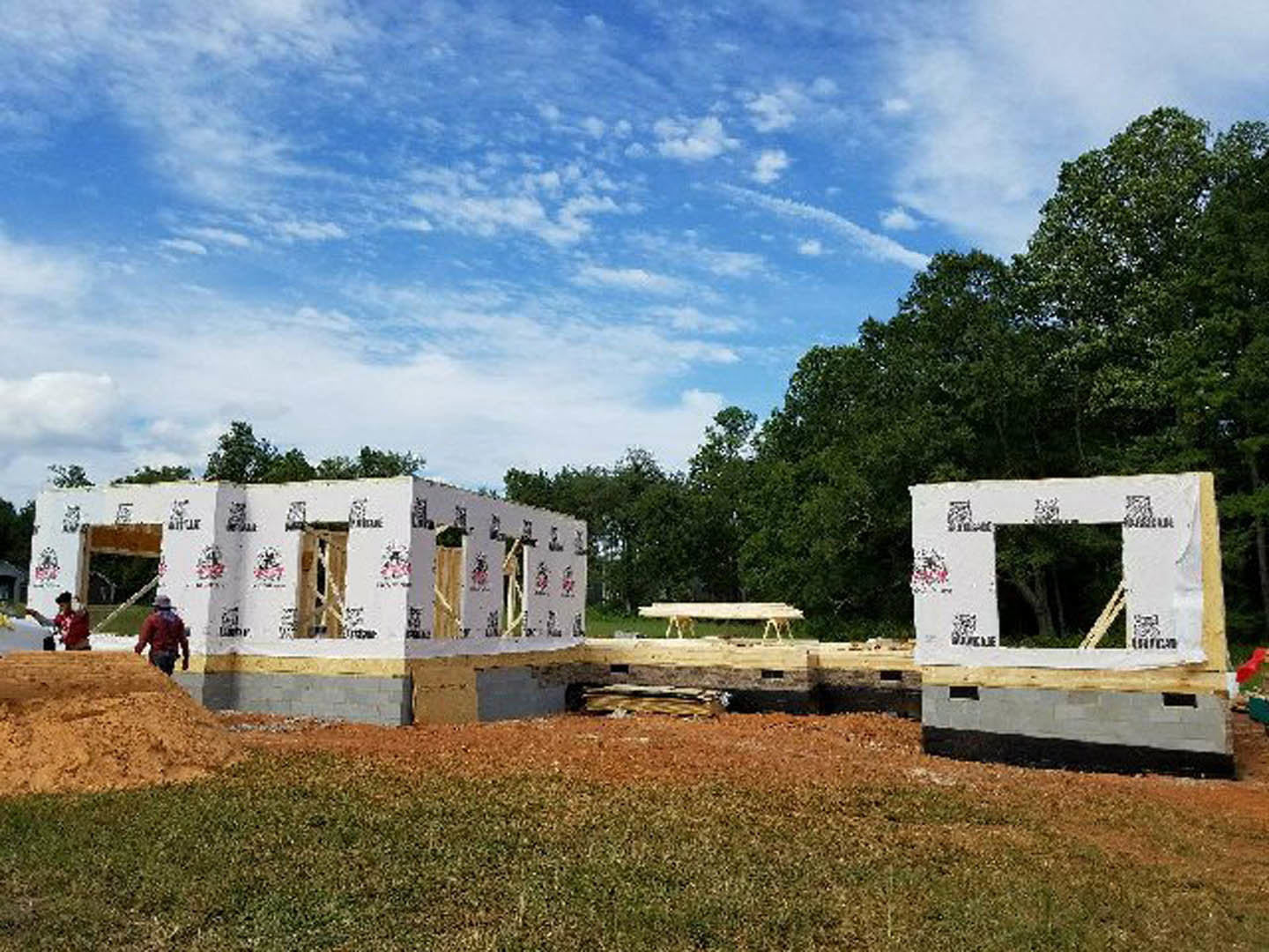 Wood-framed house under construction with exposed beams, white window frame taped in place, grassy field in foreground, cloudy sky overhead, two workers blurred in motion