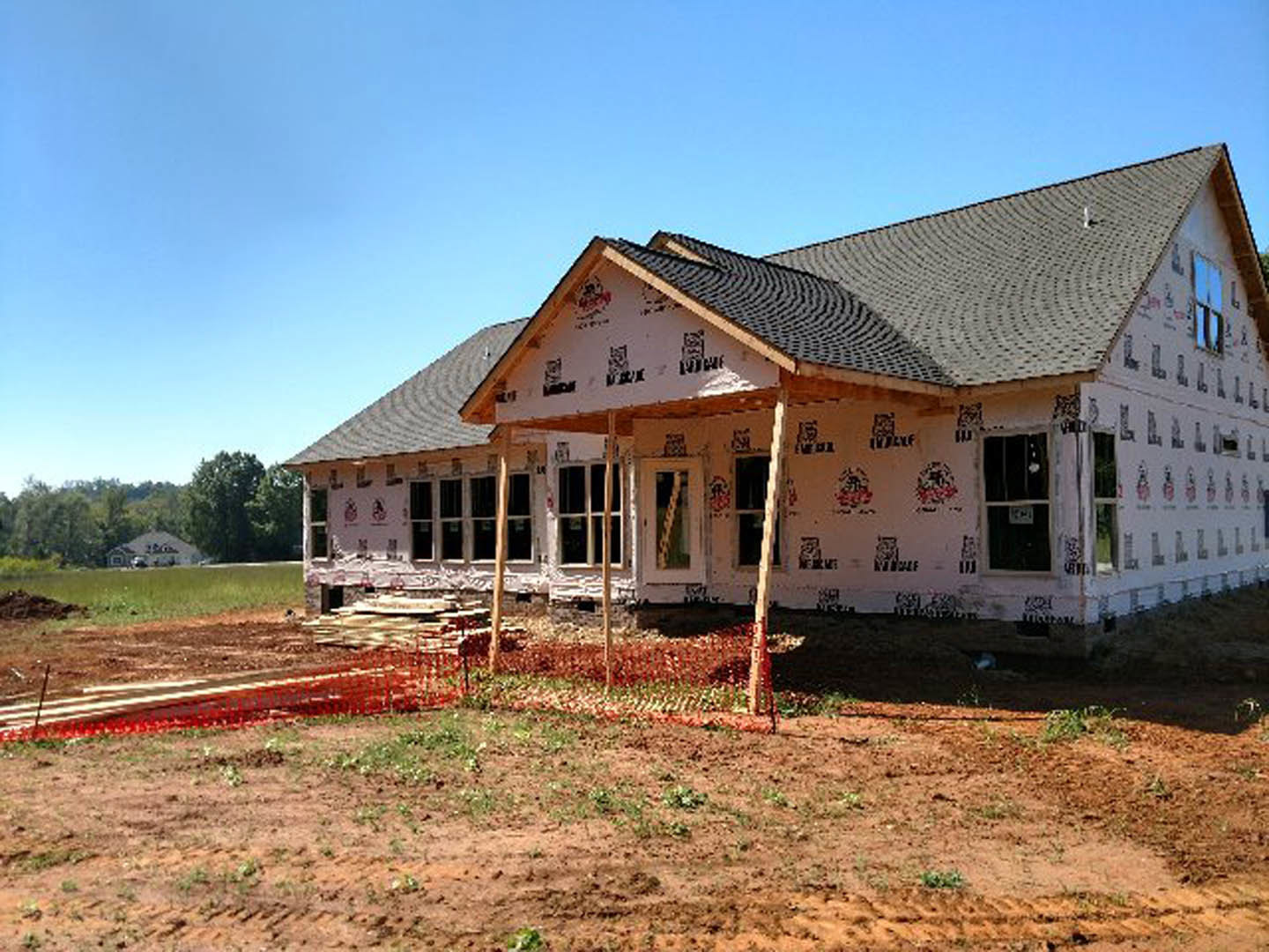 Partially built house with exposed framing, red temporary fence, covered porch, grassy yard, and surrounding trees
