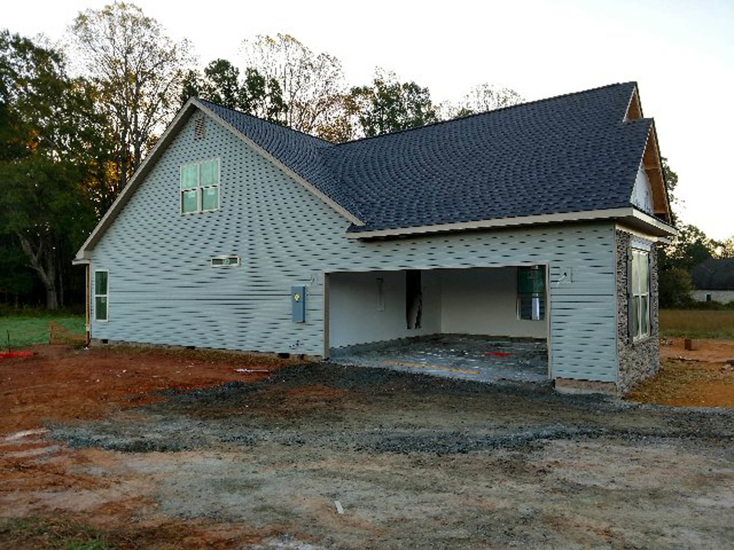 Two-story custom home with attached garage, white siding, gray shingle roof, and large windows; gravel driveway and mature trees in the yard