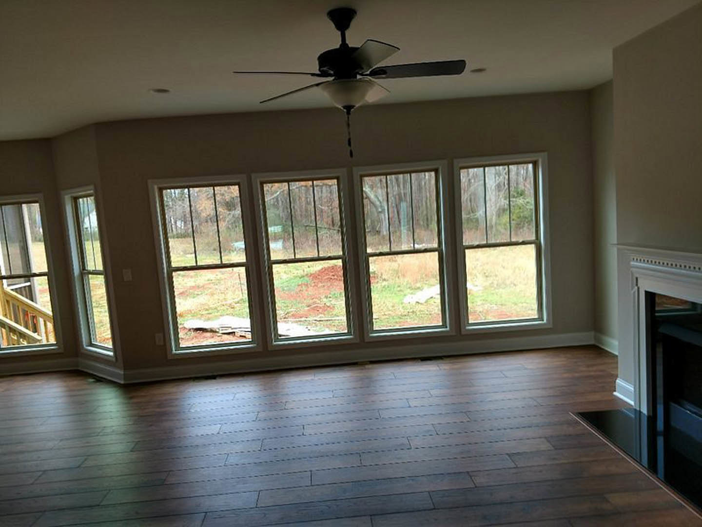 Open living area with hardwood flooring, white walls, ceiling fan with integrated light, large windows, black wall-mounted light fixture, and wooden railing.