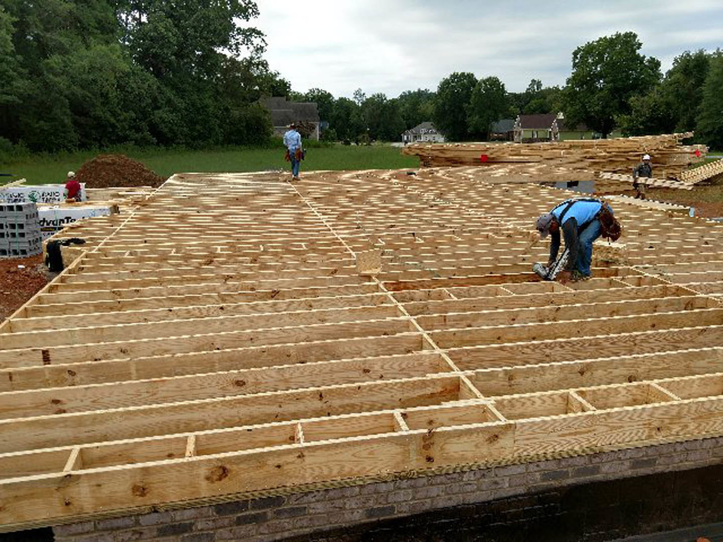 Crew installing roofing materials on a custom home, surrounded by trees and clear sky, with workers in blue shirts and jeans actively engaged on the roof.