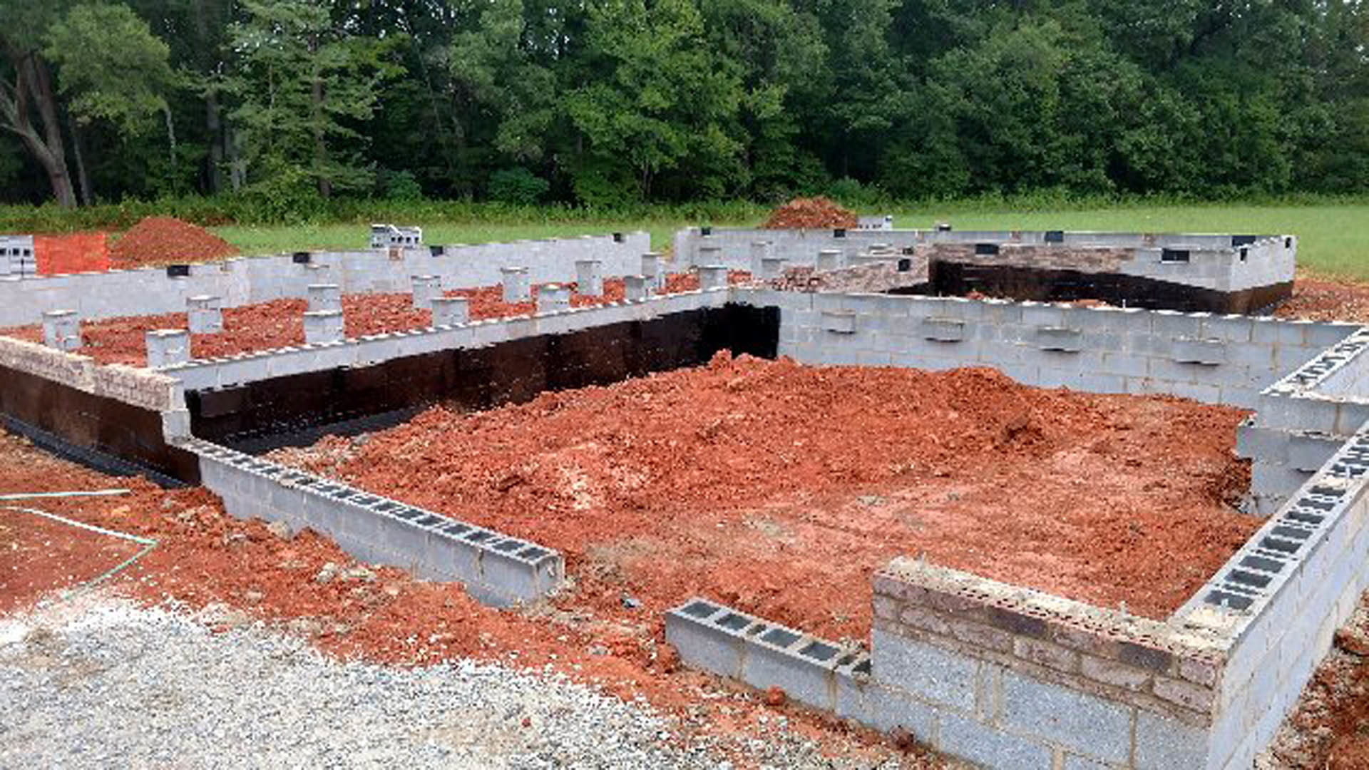 Concrete house foundation surrounded by red soil, sparse grass, and trees in the background