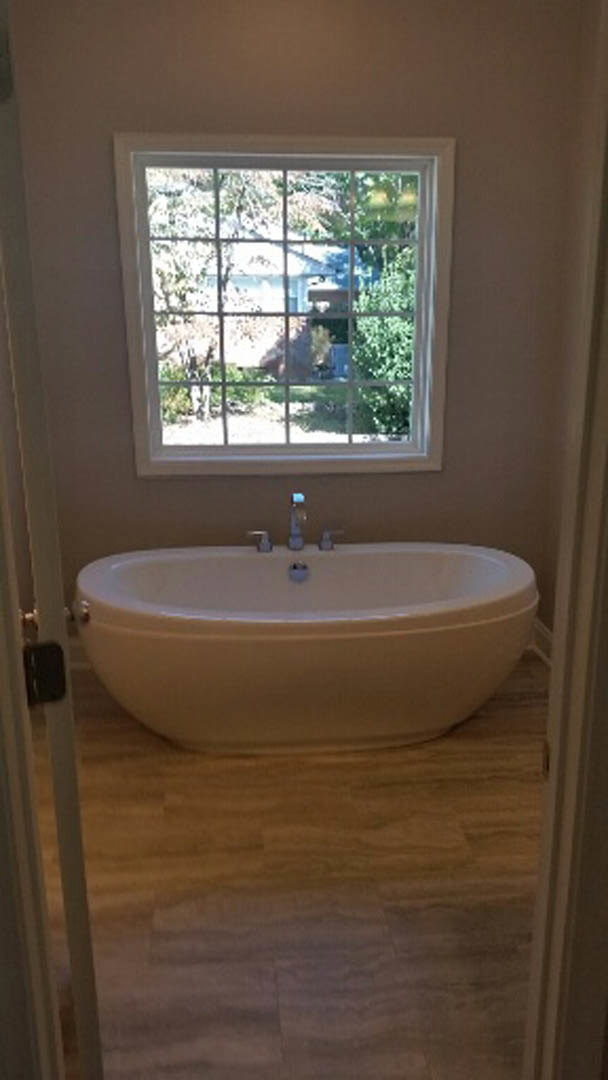 Freestanding white bathtub with chrome faucet beside large window, surrounded by light gray tile walls and floor