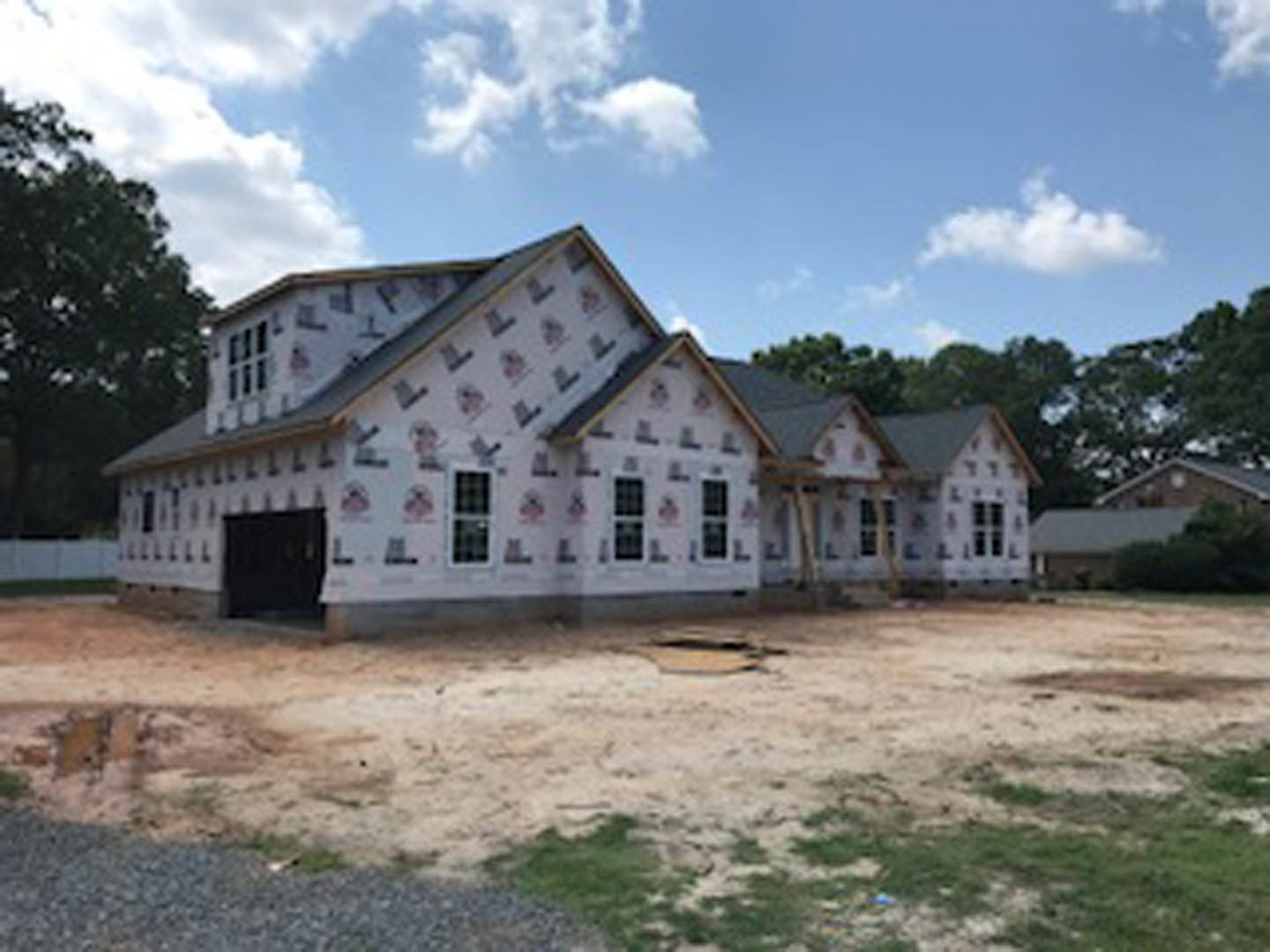 Two-story house under construction with exposed framing, dirt yard, surrounding trees, white-framed windows, attached garage, and blue sky with scattered clouds