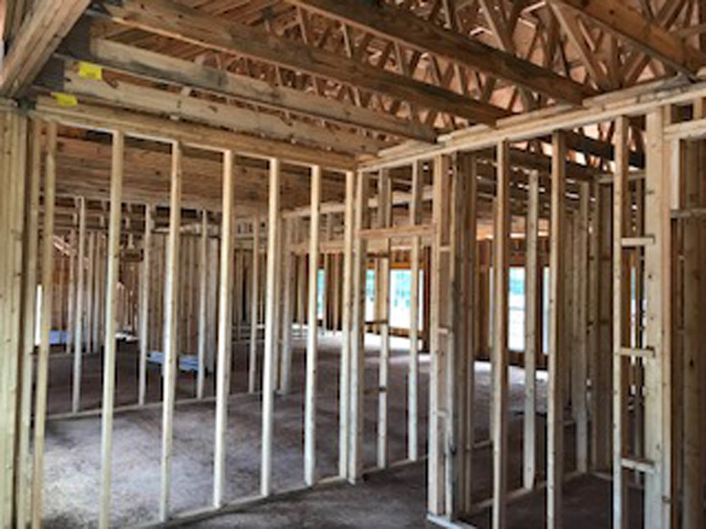 Exposed wood framing and beams with white support pillars inside an unfinished residential room, yellow construction material visible in background