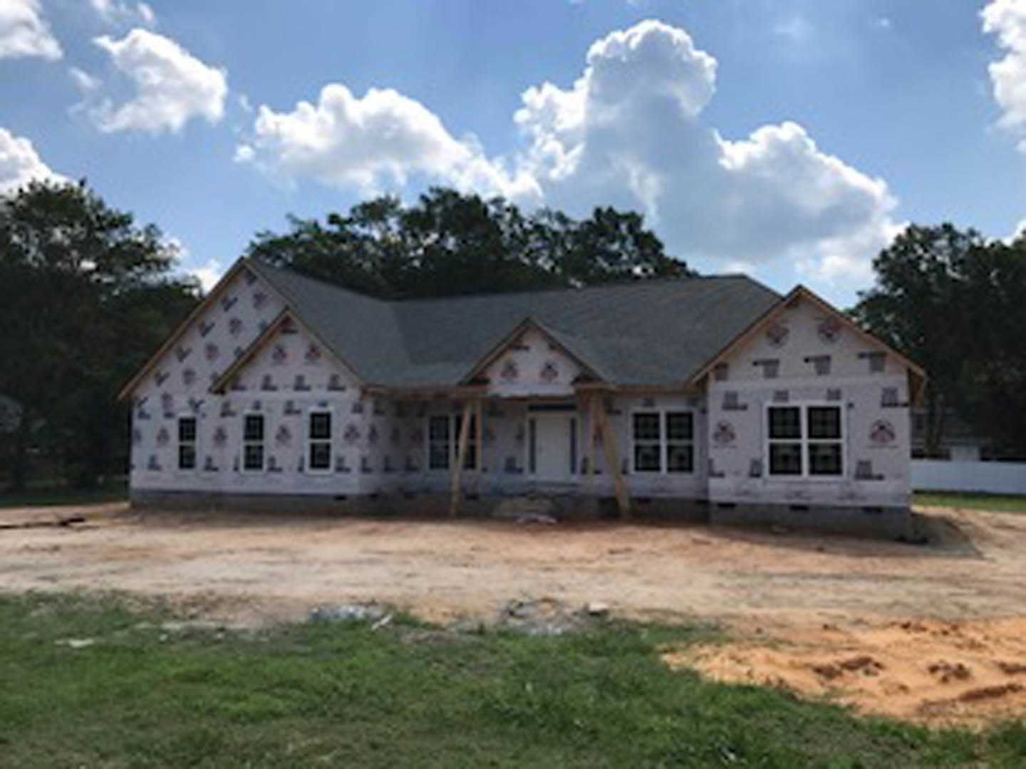 Partially built house with exposed framing, black-trimmed white door, grassy yard with patches of dirt, large windows, surrounded by trees under a blue sky with scattered clouds