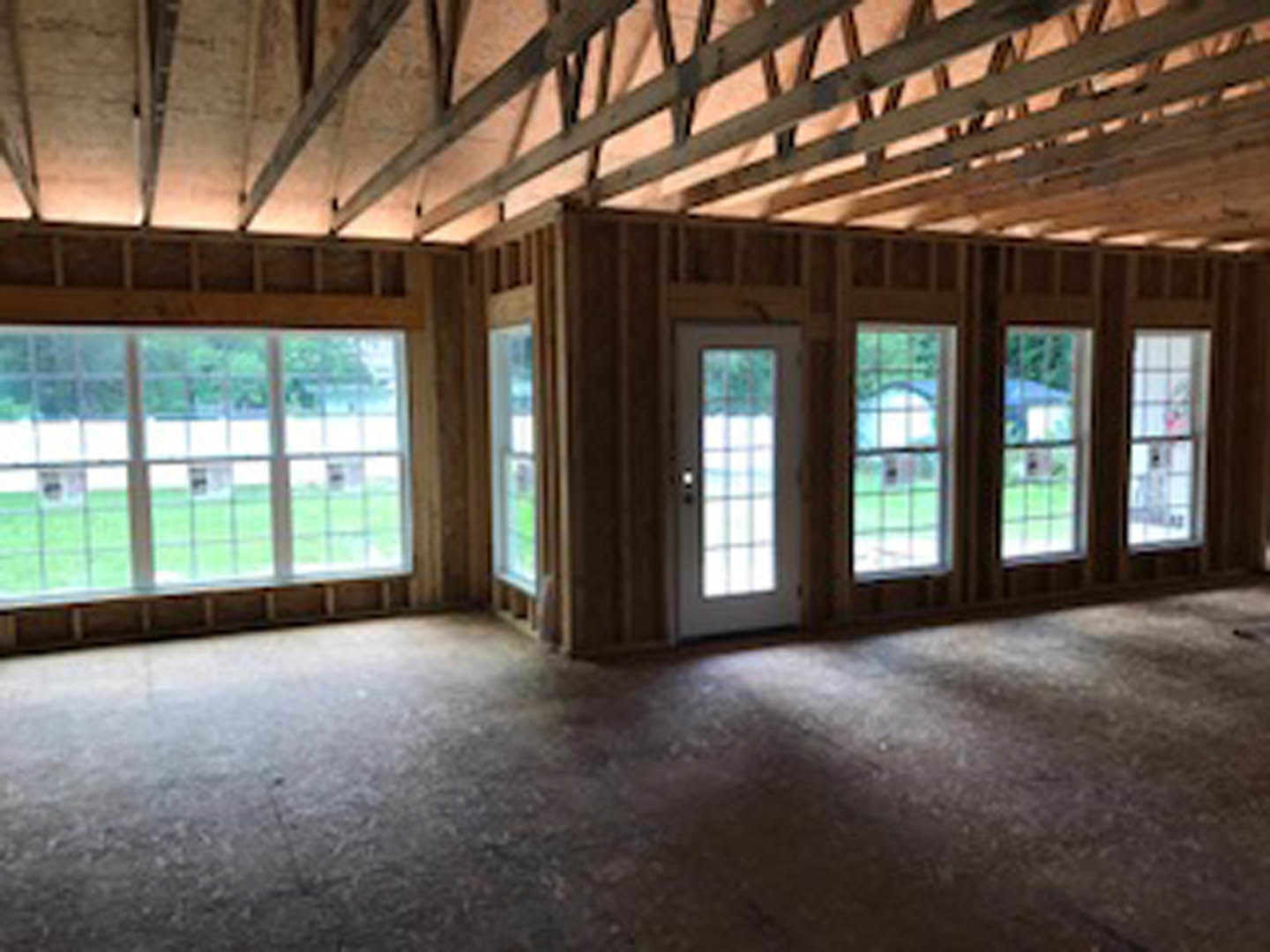 Spacious room featuring large multi-pane windows, exposed concrete flooring, white ceiling with visible beams, and a door opening to a green field.