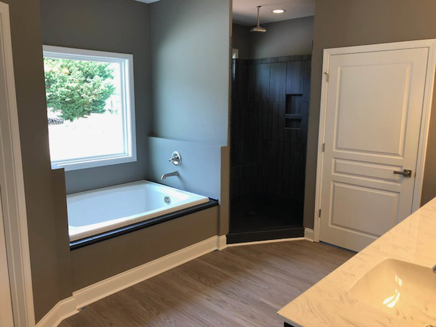 Freestanding white bathtub with chrome faucet beside a white vanity sink, light gray tile flooring, white paneled door with silver handle, frosted window, modern wall sconce, and