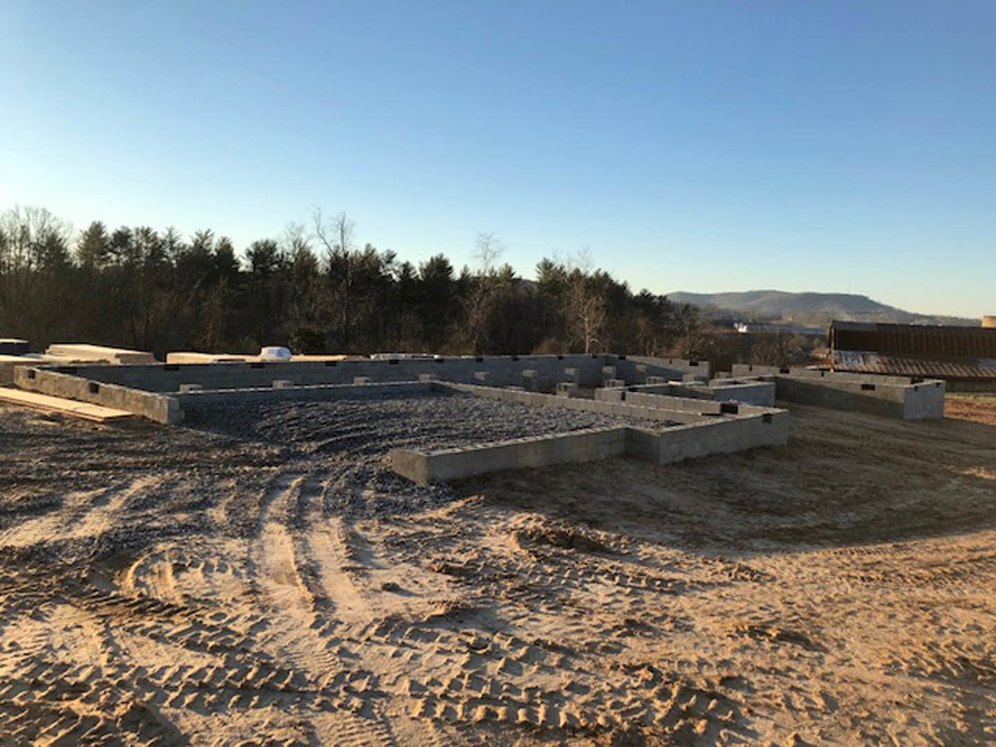Dirt construction site with tire tracks, surrounded by trees under a clear blue sky