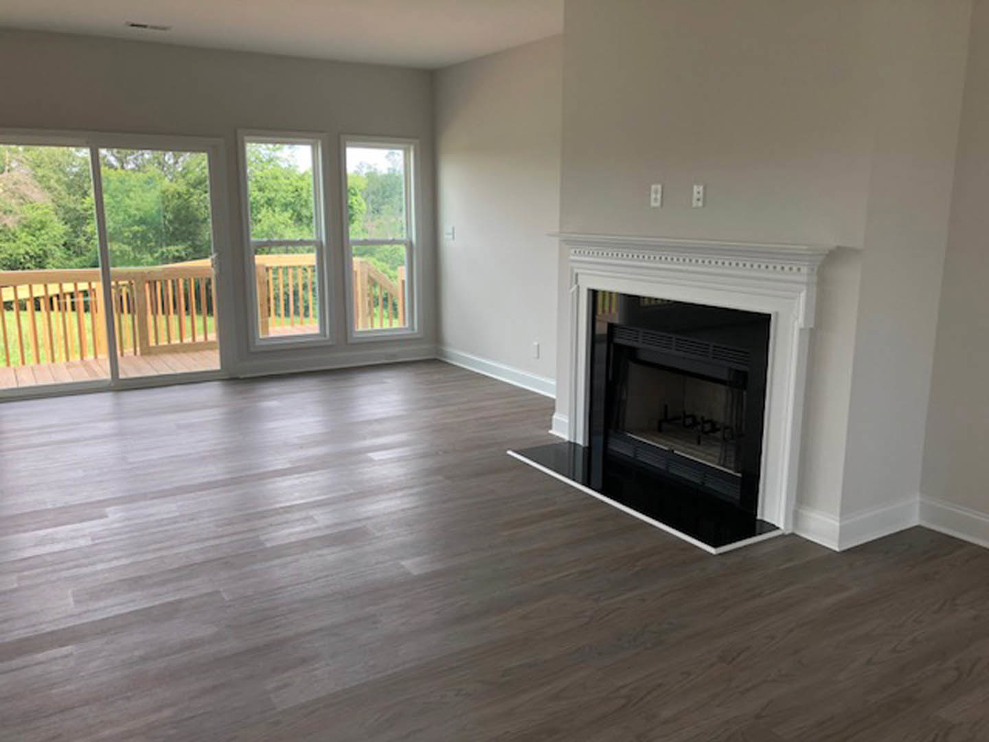 White-framed fireplace set against a light wall, wood flooring with a white trim, large window featuring a wooden railing, deck and patio visible outside