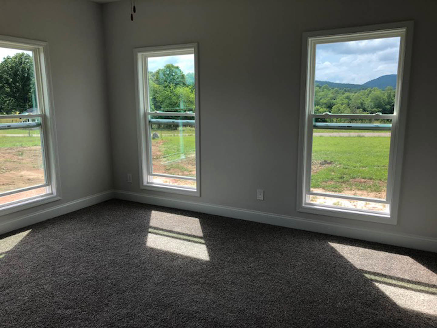 Carpeted room with two large windows, sunlight streaming onto the floor, view of grassy field and distant mountains