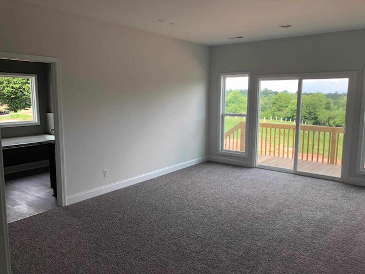 Carpeted room with large windows, wooden deck and balcony featuring wood railing, overlooking green field and trees