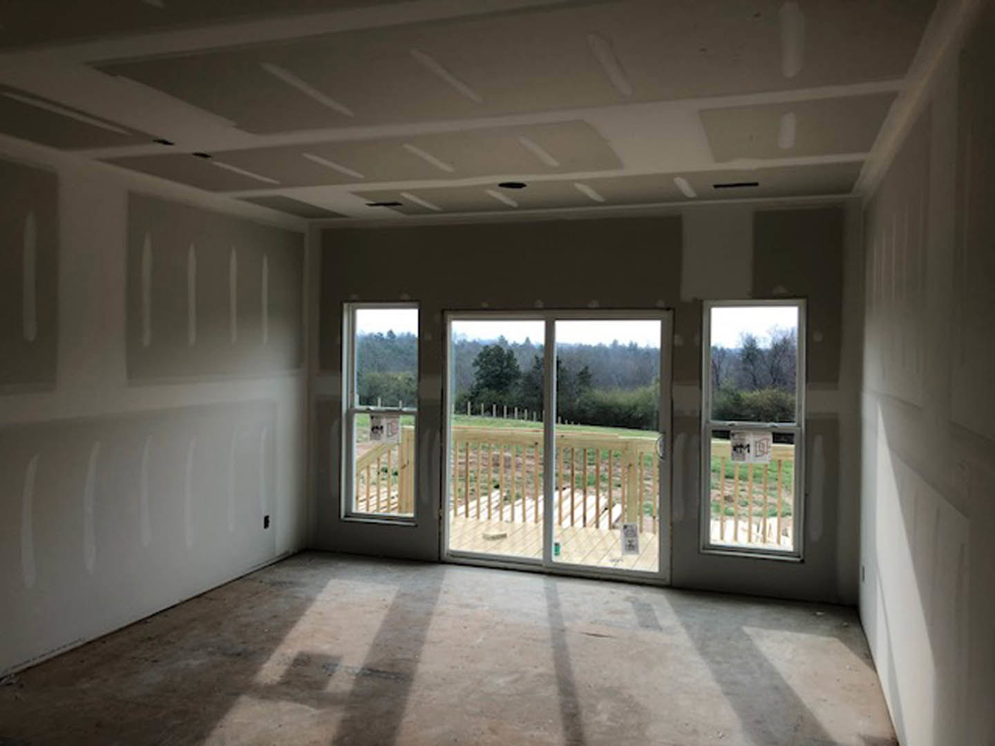 Bright room with large sliding glass door opening to deck, view of grassy yard and trees, sunlight casting shadow on light wood floor, white walls, ceiling fixtures visible