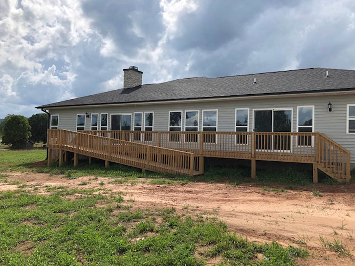 Wooden deck with railing attached to a house featuring a covered porch, brick chimney, and multiple windows under a gabled roof, surrounded by grass and open sky.