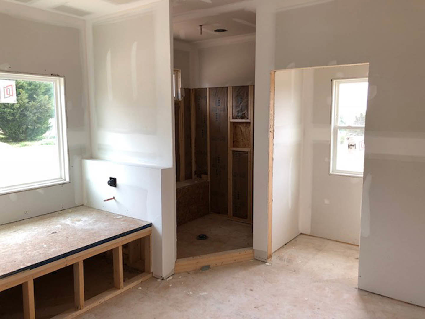 Walk-in shower with glass enclosure, white tile walls, window with white frame, wooden floor featuring circular cutouts, built-in shelf, recessed ceiling light, partial view of