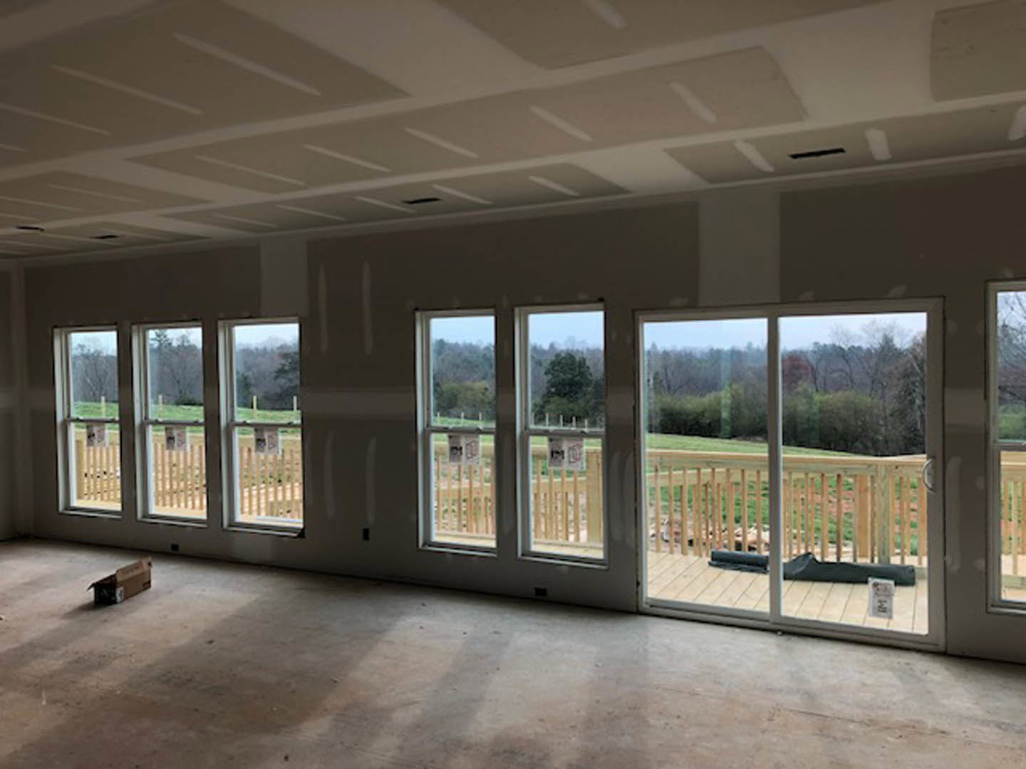 Open room with large windows, concrete floor, sliding glass door leading to deck, black bag on tile floor, brown box with black label near glass door.