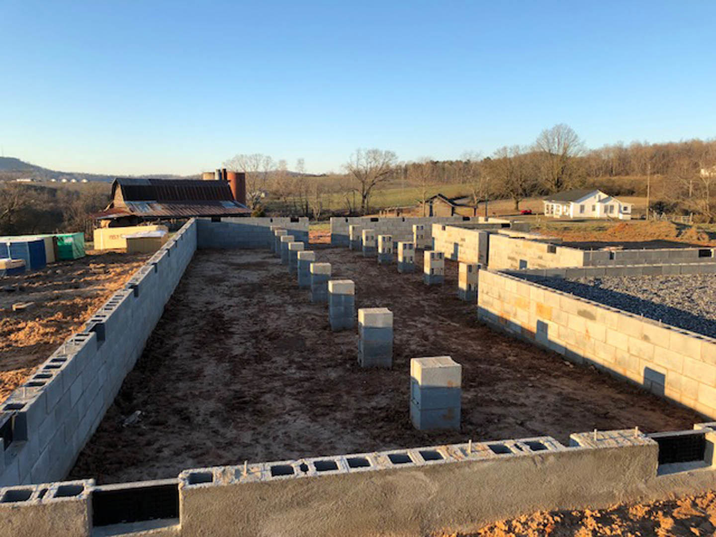 Concrete block foundation with square window openings at a residential construction site, surrounded by dirt ground and trees under a clear sky