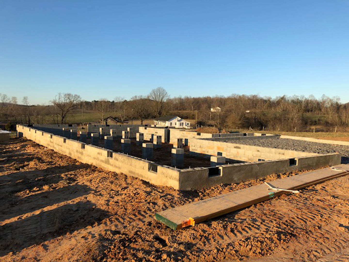 Dirt construction site with tire tracks, concrete foundation walls, wooden plank on ground, partially built house in foreground, completed house and trees in background under blue